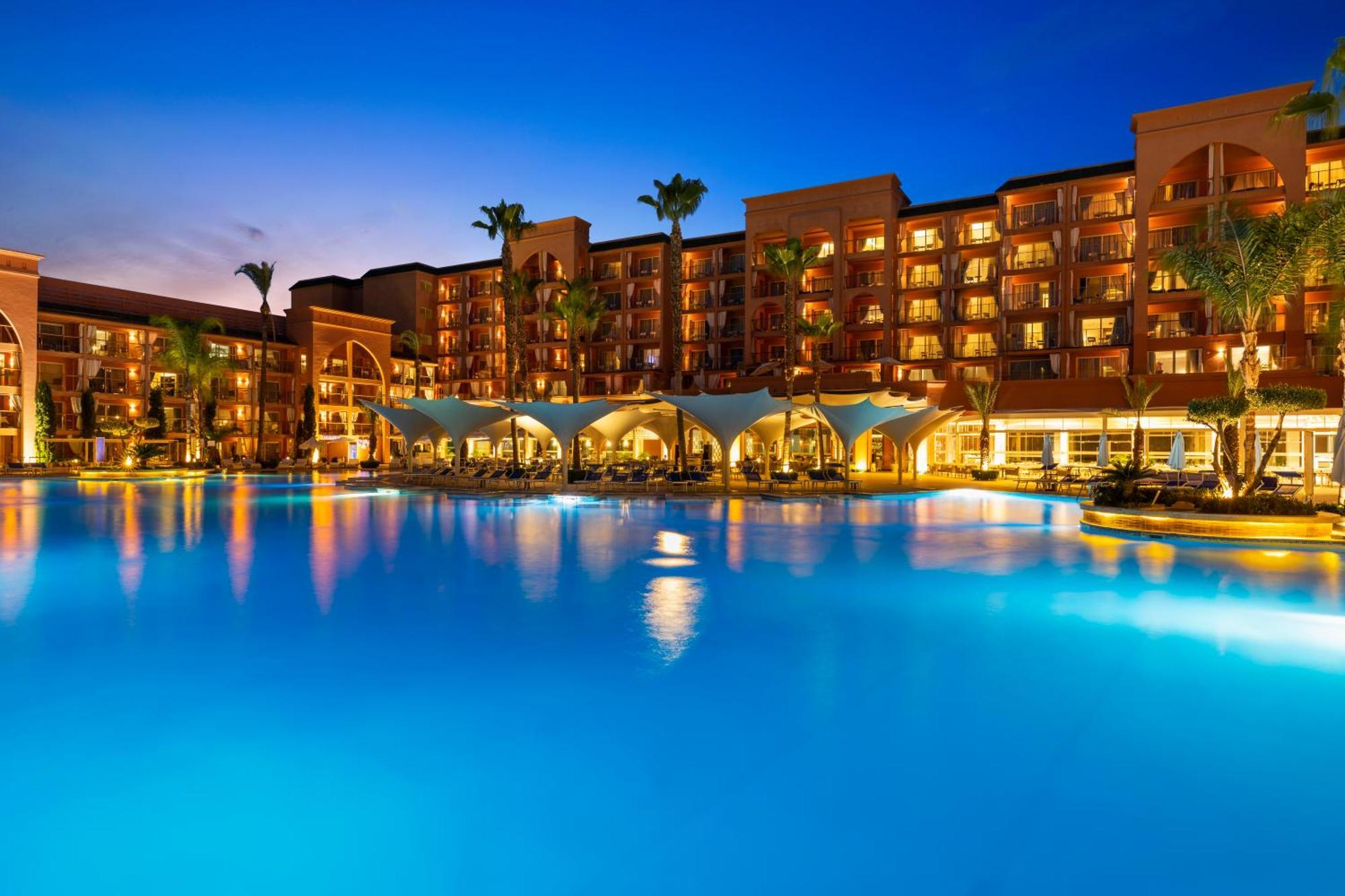 Savoy Le Grand Hotel Marrakech Marrakesh Exterior foto The photo depicts a luxurious resort setting during twilight. There is a large, illuminated swimming pool in the foreground with a vibrant blue hue, reflecting the evening sky. Surrounding the pool are several palm trees, adding a tropical feel to th