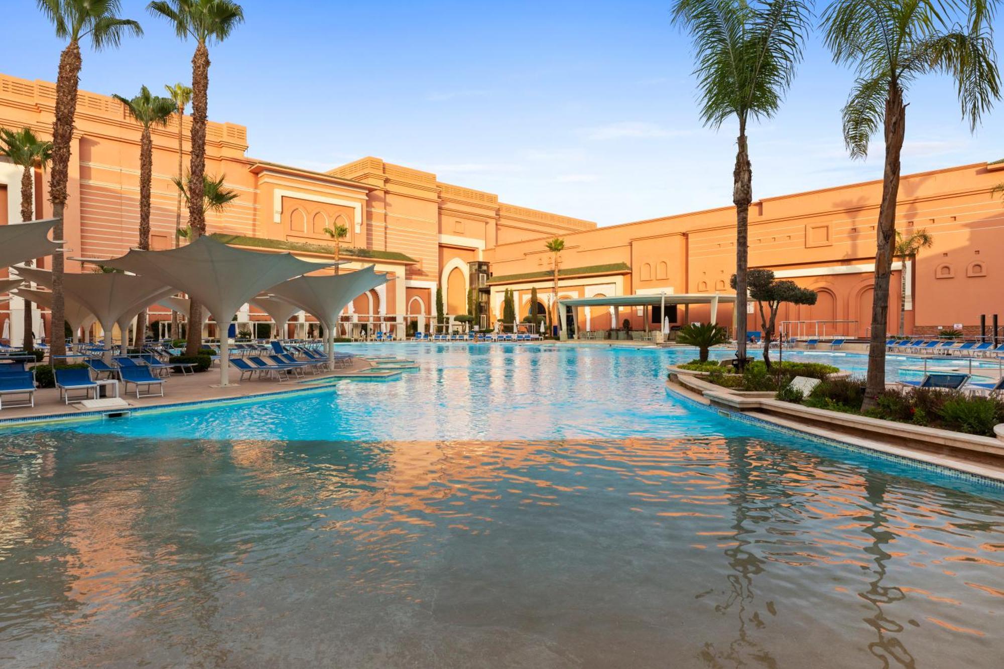 Savoy Le Grand Hotel Marrakech Marrakesh Exterior foto The photo depicts a luxurious pool area surrounded by palm trees and a beautiful architectural backdrop. The water is clear and inviting, with lounge chairs neatly arranged along the poolside. The design features a blend of modern and classical eleme