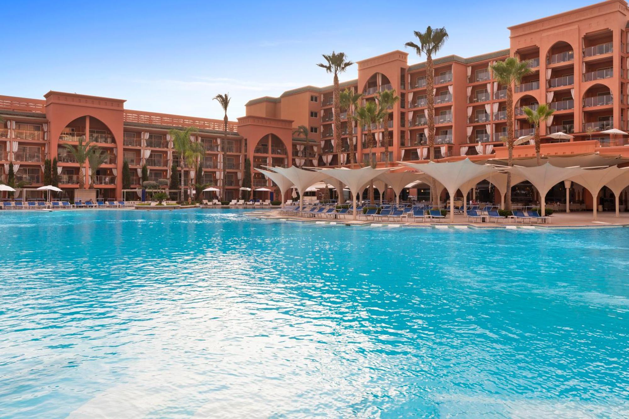 Savoy Le Grand Hotel Marrakech Marrakesh Exterior foto The photo shows a luxurious hotel or resort with a large, inviting swimming pool in the foreground. The pool features a vibrant blue color and is surrounded by comfortable lounge chairs and shaded areas. In the background, there are multiple building