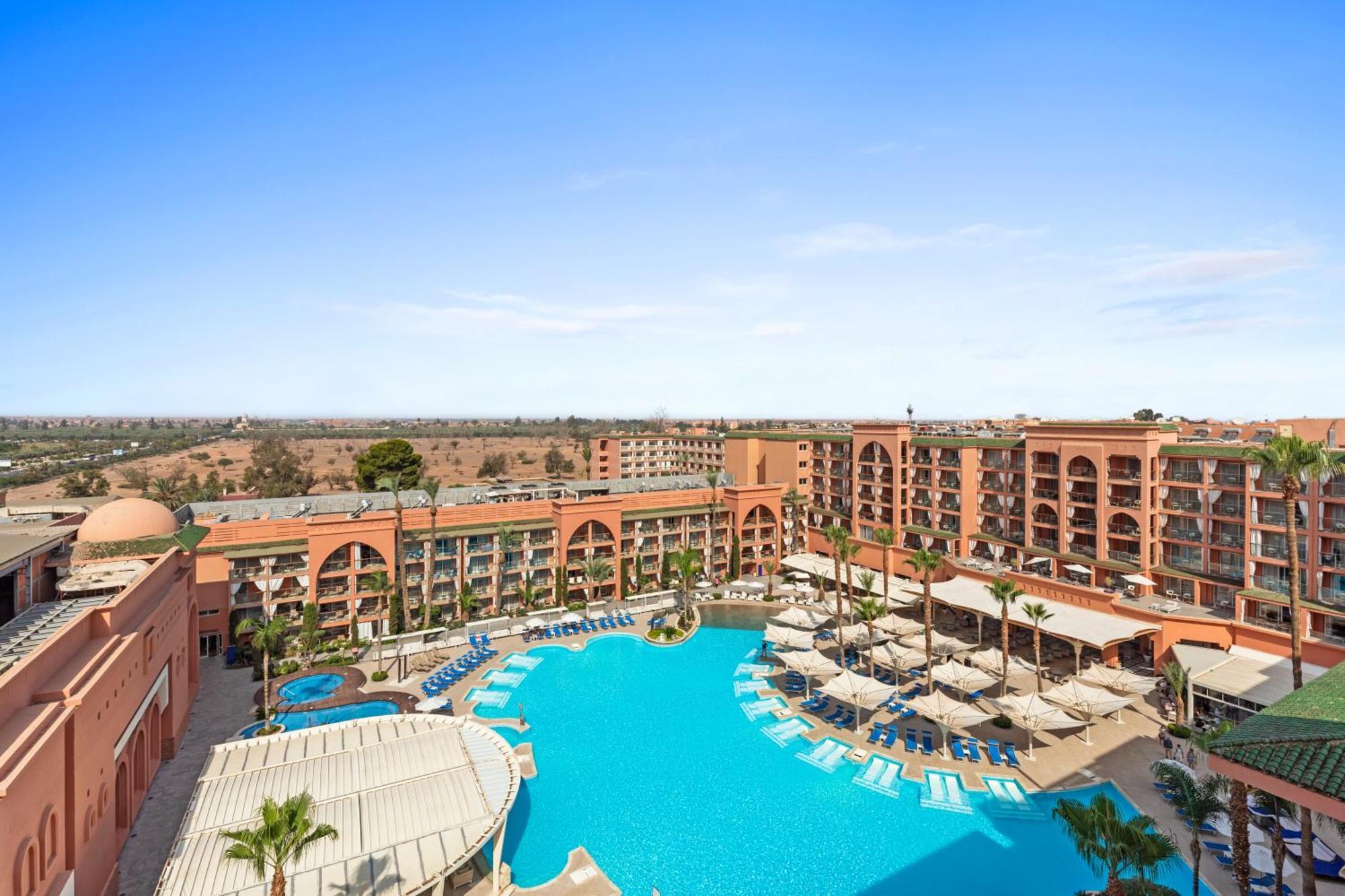 Savoy Le Grand Hotel Marrakech Marrakesh Exterior foto The image shows a resort with a large swimming pool in the foreground, surrounded by lounge chairs and umbrellas. The pool area is adorned with palm trees, contributing to a tropical vibe. In the background, there is a multi-story building featuring 