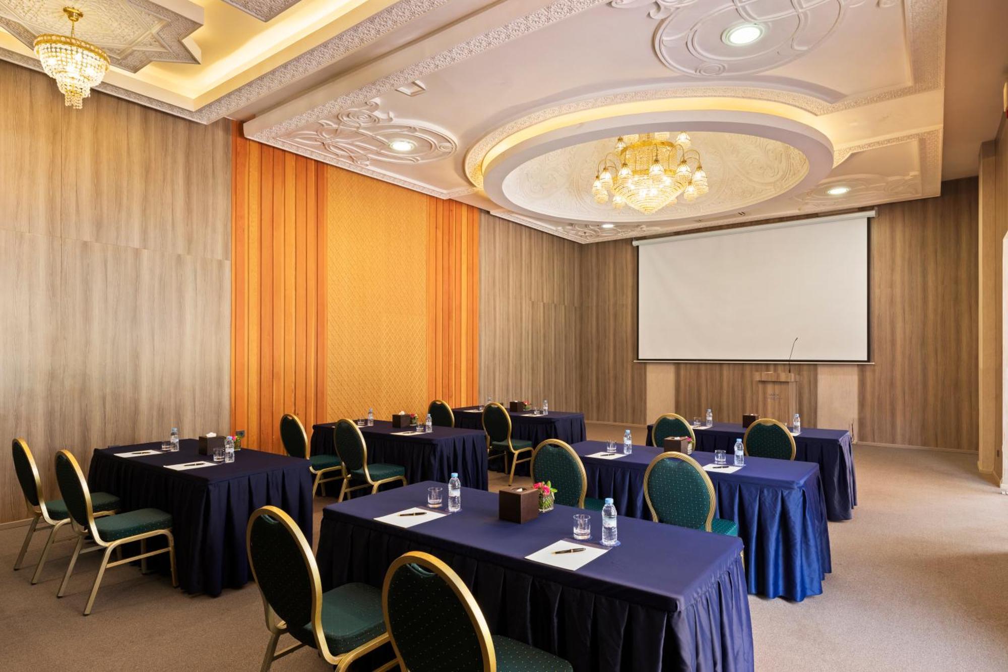 Savoy Le Grand Hotel Marrakech Marrakesh Exterior foto The photo shows a conference or meeting room. It features several tables arranged in a classroom or seminar style, each covered with a dark blue tablecloth. There are green chairs positioned around the tables. On the tables, there are water bottles a