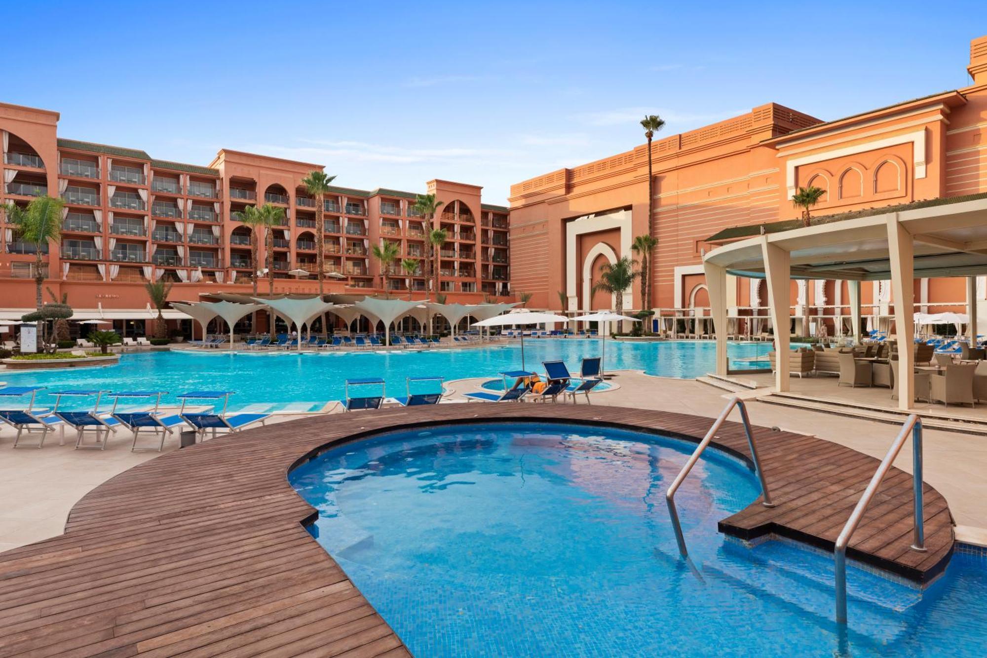 Savoy Le Grand Hotel Marrakech Marrakesh Exterior foto The photo showcases a vibrant resort with a large swimming pool and a smaller jacuzzi area. The pool area is surrounded by lounge chairs, providing space for relaxation. In the background, there are several multi-story buildings designed with a Moroc