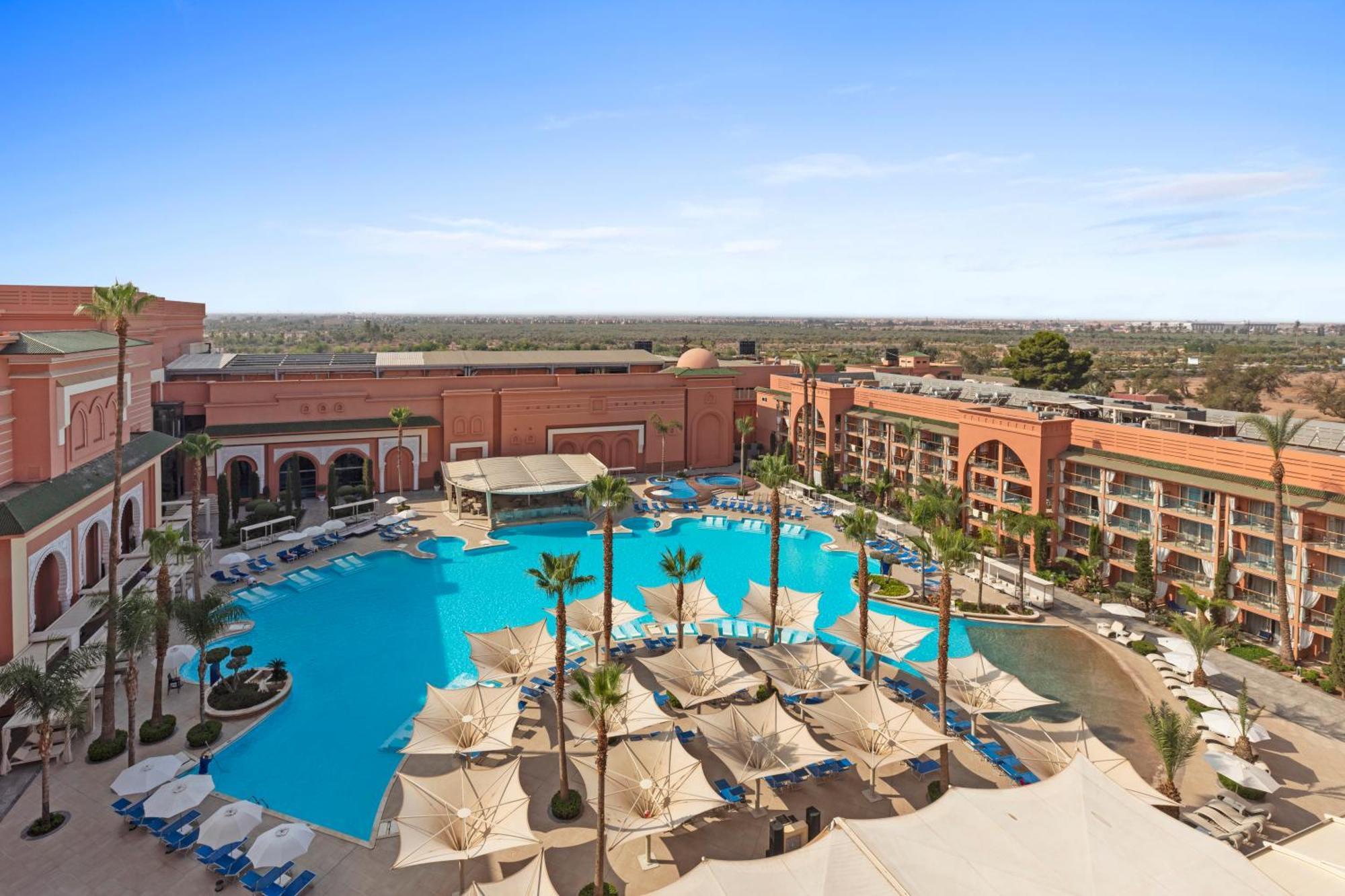 Savoy Le Grand Hotel Marrakech Marrakesh Exterior foto The photo shows a luxurious hotel resort featuring a large swimming pool at the center. Surrounding the pool are several palm trees and sun loungers, suggesting a relaxing outdoor atmosphere. There are shaded cabanas or umbrellas providing cover for 