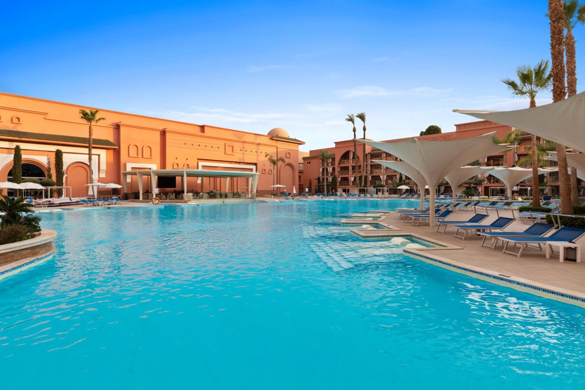 Savoy Le Grand Hotel Marrakech Marrakesh Exterior foto The photo shows a large outdoor swimming pool with clear, turquoise water. Surrounding the pool are several lounge chairs, some of which are sheltered by large umbrellas. In the background, there are buildings with a distinctly warm-colored architect