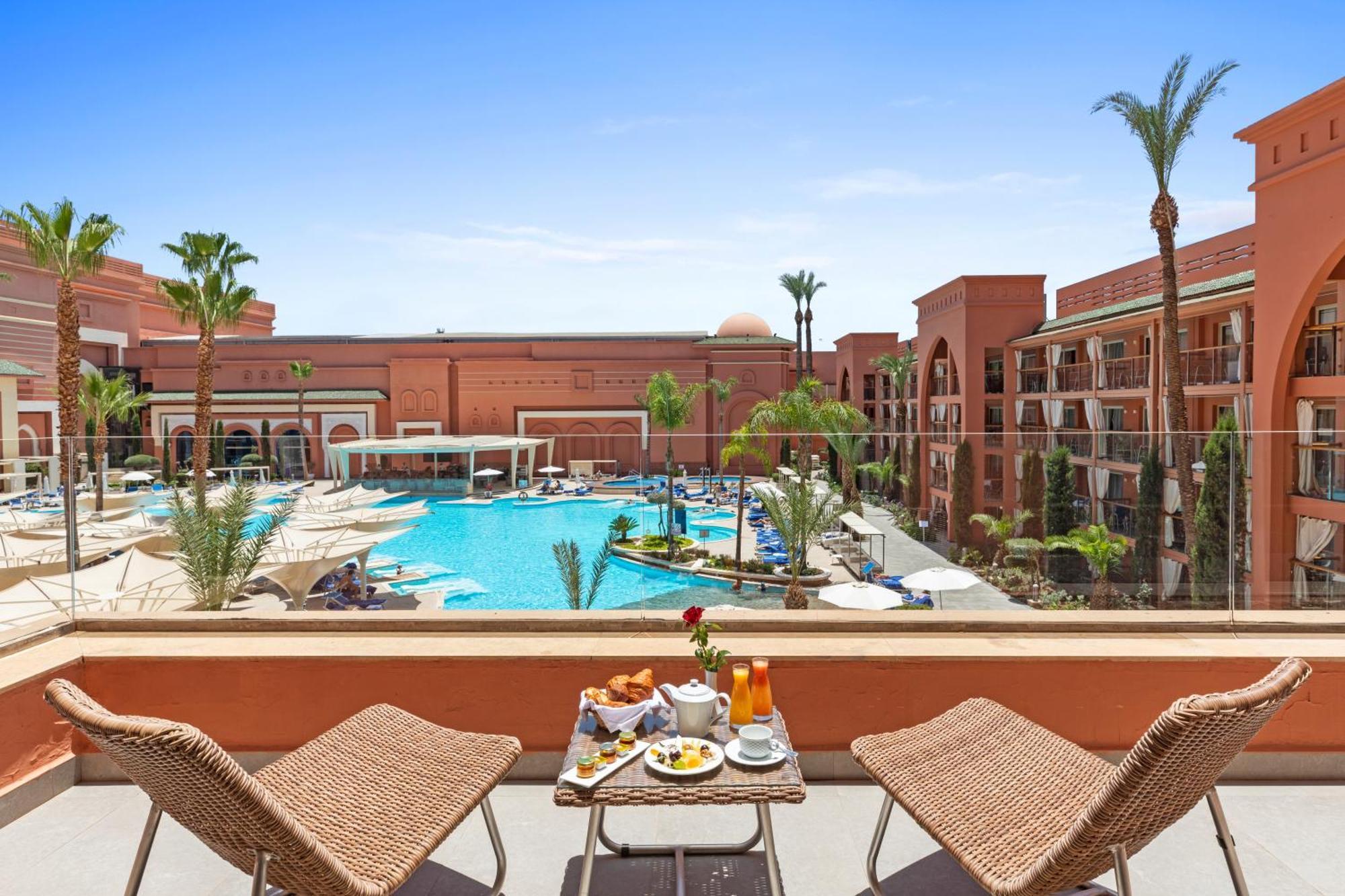 Savoy Le Grand Hotel Marrakech Marrakesh Exterior foto The photo shows a picturesque view from a balcony overlooking a hotel pool area. In the foreground, there are two wicker chairs and a small table set with breakfast items, including a vase with a flower, a juice glass, and some food. In the backgroun