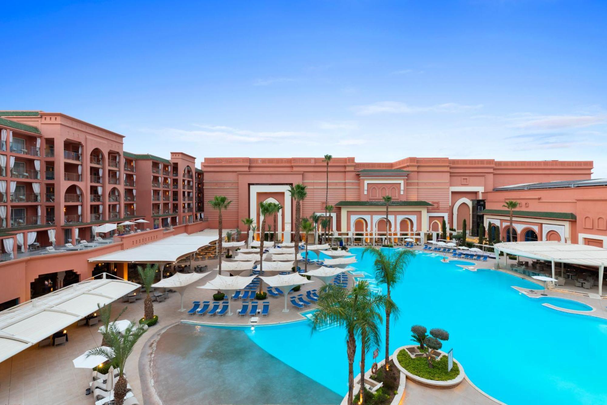 Savoy Le Grand Hotel Marrakech Marrakesh Exterior foto The photo depicts a luxurious resort featuring an expansive pool area. The pool is a vibrant blue and surrounded by palm trees and lounge chairs. In the background, there are elegantly designed buildings with a reddish stone facade, characteristic of