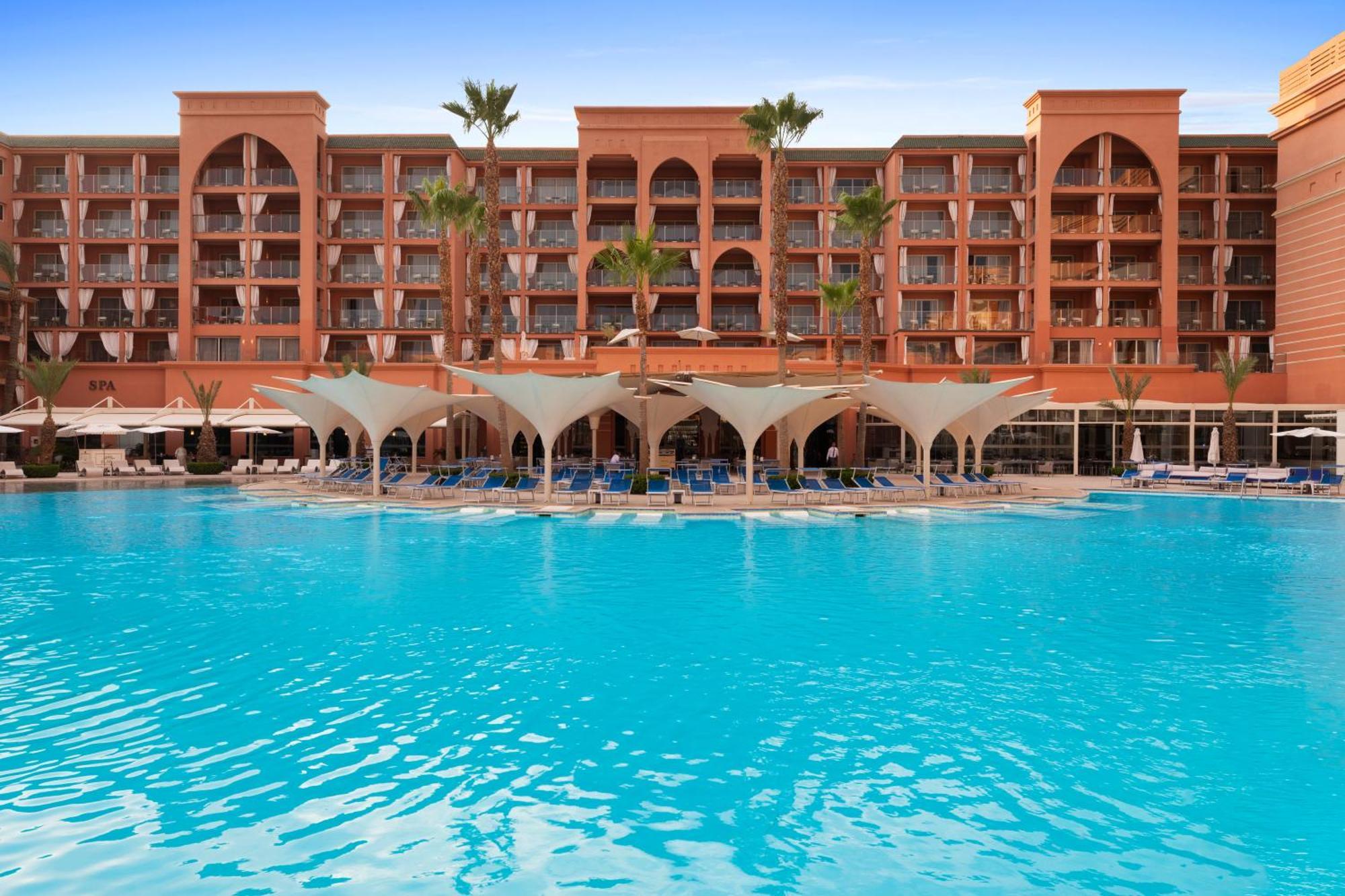 Savoy Le Grand Hotel Marrakech Marrakesh Exterior foto The photo shows a beautiful hotel with an architectural style typical of Moroccan design, characterized by its red-orange facade and arched windows. In the foreground, there is a large swimming pool with crystal-clear blue water. The pool area is sha