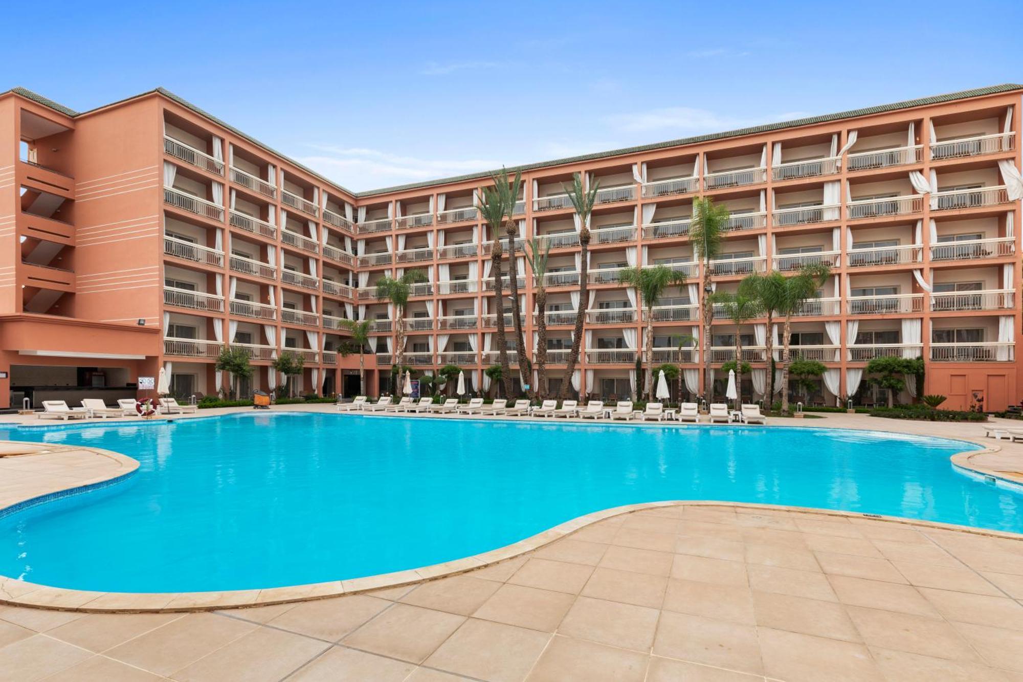 Savoy Le Grand Hotel Marrakech Marrakesh Exterior foto The photo shows a large swimming pool surrounded by a paved area with lounge chairs. In the background, there is a multi-story building with a pink exterior featuring multiple balconies. The setting appears to be sunny, and there are palm trees nearb