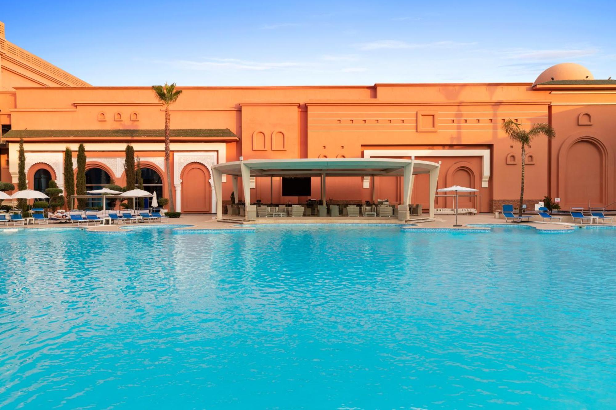 Savoy Le Grand Hotel Marrakech Marrakesh Exterior foto The photo depicts a luxurious outdoor pool area of a resort or hotel. In the foreground, there is a large, inviting swimming pool with bright turquoise water. Surrounding the pool are comfortable lounge chairs and umbrellas, suggesting a relaxing atm