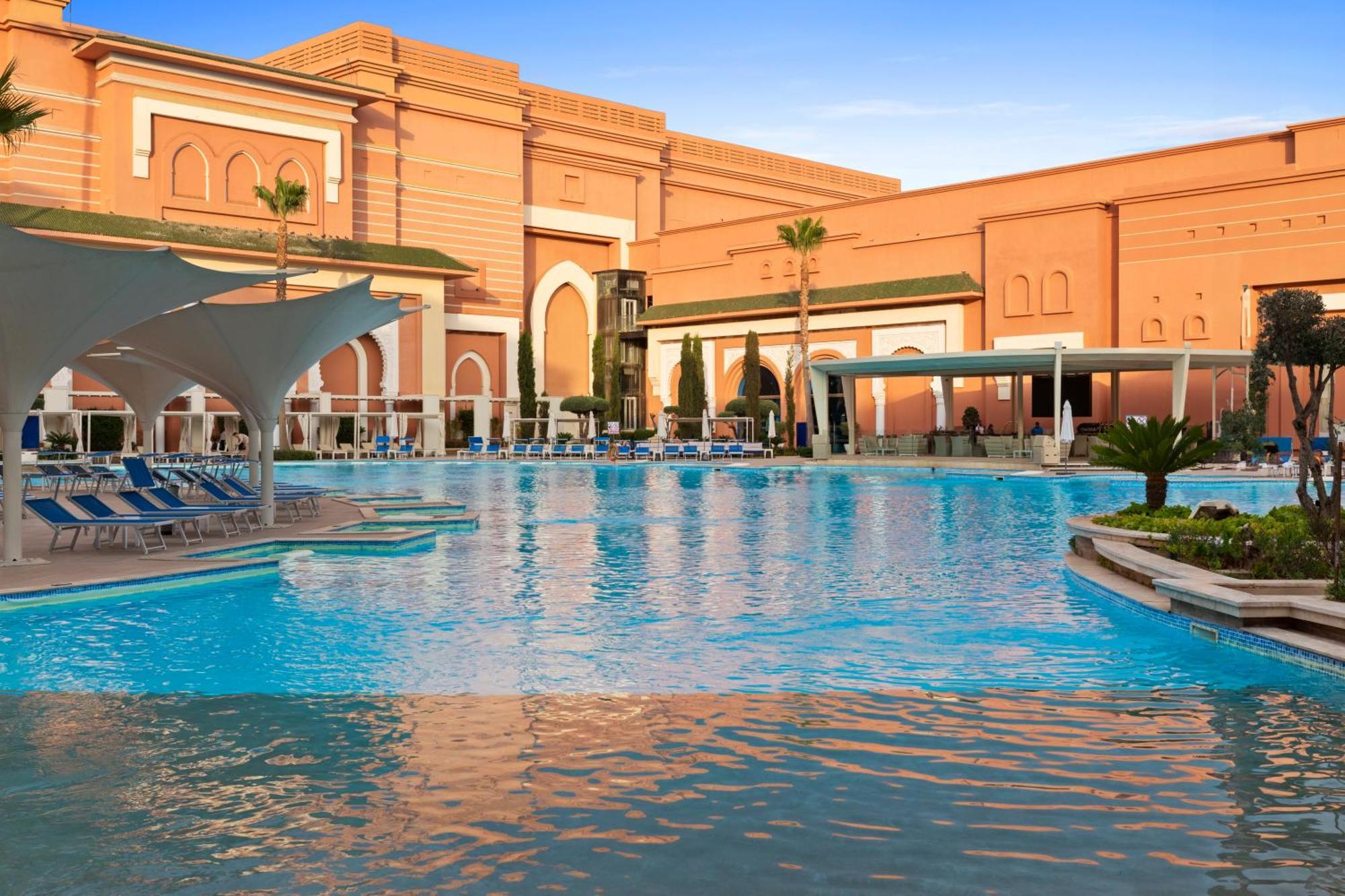 Savoy Le Grand Hotel Marrakech Marrakesh Exterior foto The photo shows a large, inviting swimming pool with clear blue water, surrounded by lounge chairs. In the background, there is an elegantly designed building with a warm-colored facade, featuring arches and decorative elements, typical of luxury res