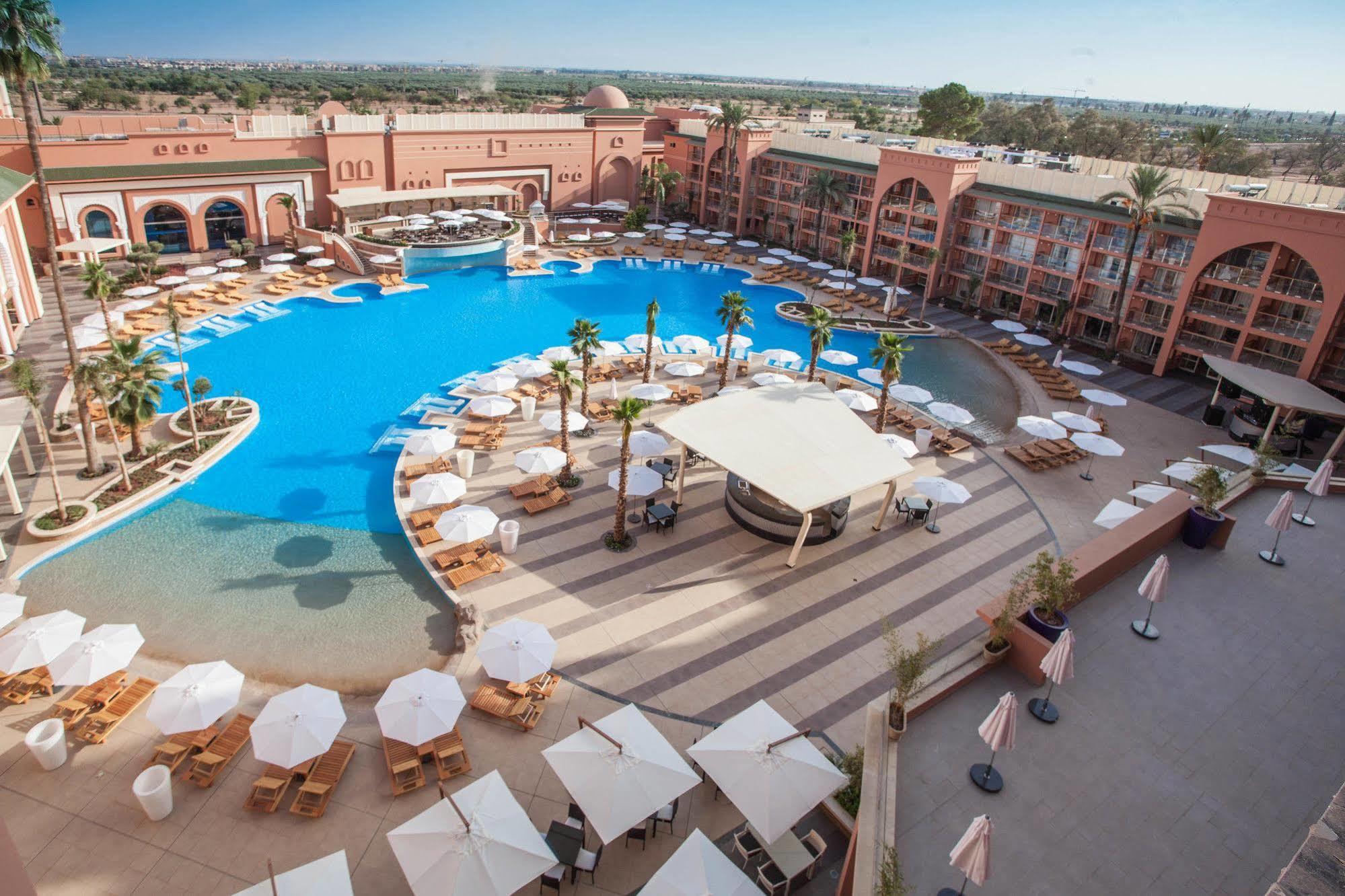 Savoy Le Grand Hotel Marrakech Marrakesh Exterior foto The photo shows a large, luxurious hotel resort with a spacious swimming pool at its center. The pool area features several lounge chairs and umbrellas arranged around the water, providing a relaxing atmosphere for guests. Surrounding the pool are la